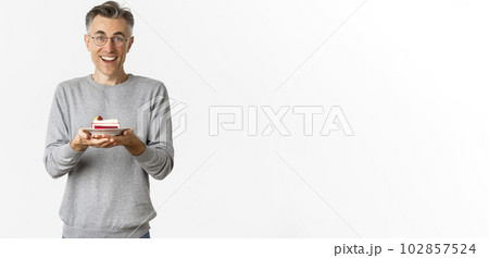 Portrait of happy handsome middle-aged man, wearing glasses and gray sweater, holding cake and looking amazed at camera, standing over white background 102857524