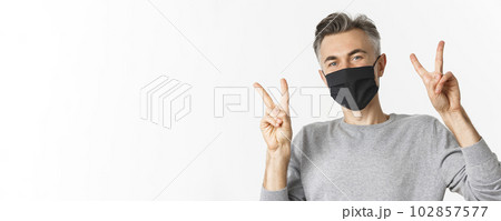 Concept of covid-19, social distancing and quarantine. Close-up of happy and cute middle-aged guy in black medical mask, showing peace signs and smiling, standing over white background Concept of covid-19, social distancing and quarantine. Close-up of happy and cute middle-aged guy in black medical mask, showing peace signs and smiling, standing over white background 102857577