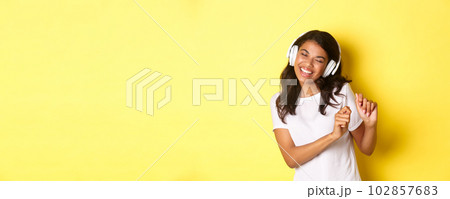 Cheerful teenage african-american girl listening to music in headphones, dancing upbeat and smiling, standing over yellow background 102857683