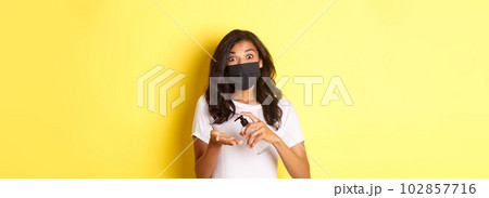 Concept of covid-19, social distancing and lifestyle. Image of african-american girl in face mask using hand sanitizer, standing over yellow background 102857716