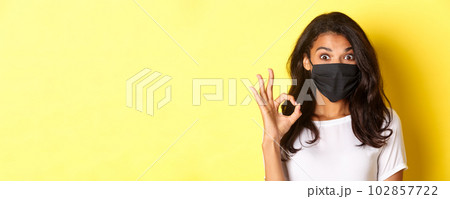 Concept of coronavirus, pandemic and lifestyle. Close-up of amazed and satisfied african-american girl, wearing black face mask, showing okay sign, like something good, yellow background 102857722