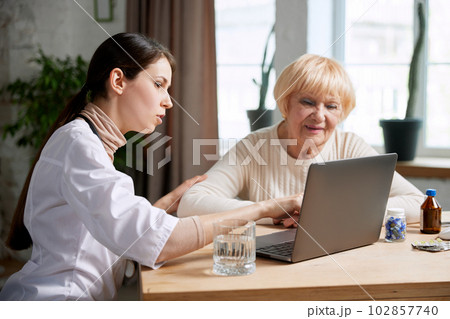 Female doctor sitting at table in office with her senior patient and talking, looking on laptop. Explaining medical results 102857740