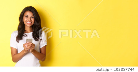 Image of modern african-american girl smiling, using mobile phone, standing over yellow background 102857744