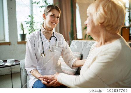 Smiling young woman, doctor giving support and hope to her elderly female patient. Woman holding hand of senior lady at home 102857765