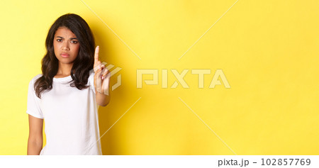Image of disappointed african-american girl telling no, shaking finger to prohibit or stop someone, disagree with person, standing over yellow background 102857769