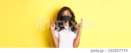 Concept of covid-19, social distancing and lifestyle. Image of excited and hopeful african-american girl, making a wish, wearing face mask and crossing fingers for good luck, over yellow background 102857774
