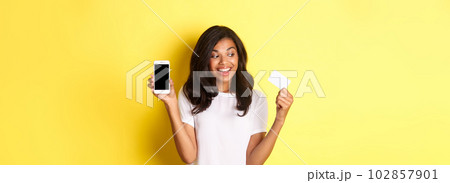 Image of excited african-american female model, showing smartphone screen and credit card, standing over yellow background Image of excited african-american female model, showing smartphone screen and credit card, standing over yellow background 102857901