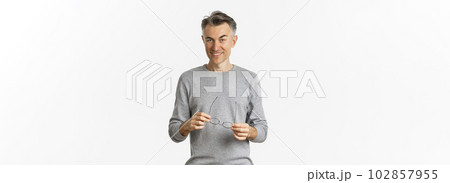 Portrait of cheeky smiling man in grey sweater, holding glasses and looking cunning at camera, standing over white background Portrait of cheeky smiling man in grey sweater, holding glasses and looking cunning at camera, standing over white background 102857955