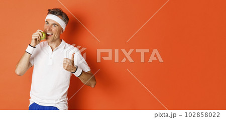 Portrait of healthy and active middle-aged man, eating an apple and showing thumbs-up, standing against orange background 102858022