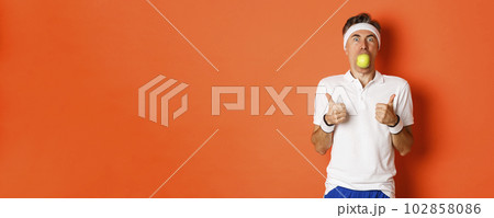 Image of funny middle-aged guy in fitness clothing, holding apple in mouth, showing thumbs-up, standing over orange background 102858086