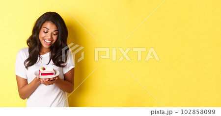 Portrait of attractive african-american woman, looking at delicious piece of cake and smiling, standing over yellow background 102858099