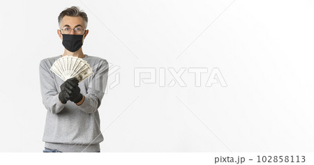 Concept of covid-19, social distancing and lifestyle. Handsome and lucky middle-aged man in glasses, medical mask and gloves, showing prize money, winning lottery, standing over white background Concept of covid-19, social distancing and lifestyle. Handsome and lucky middle-aged man in glasses, medical mask and gloves, showing prize money, winning lottery, standing over white background 102858113
