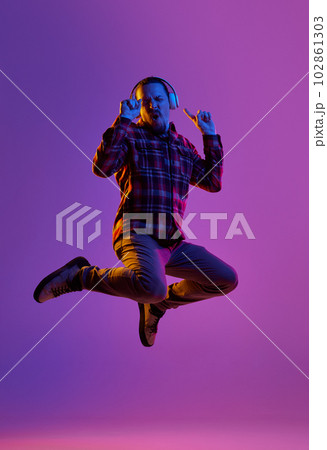 Full-length portrait of emotional man in casual clothes, listening to music in headphones and jumping against purple studio background in neon light Full-length portrait of emotional man in casual clothes, listening to music in headphones and jumping against purple studio background in neon light 102861303