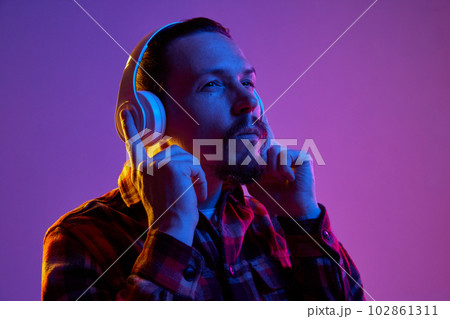 Portrait of young man in checkered shirt listening to music in headphones against purple studio background in neon light 102861311
