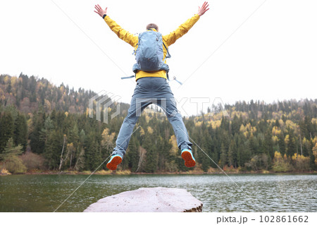 Happy male traveler with backpack jumping near forest lake back view 102861662