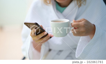 Woman in white bathrobe drinking coffee from cup and holding mobile phone at home closeup Woman in white bathrobe drinking coffee from cup and holding mobile phone at home closeup 102861781