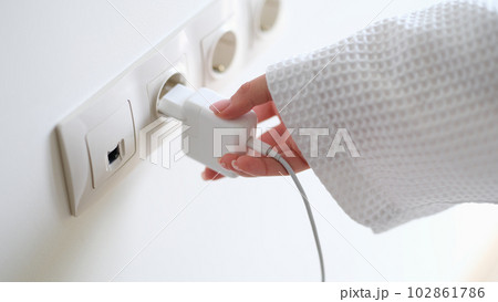 Woman in white bathrobe inserting plug into outlet at home with hand closeup 102861786