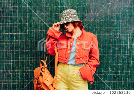 Hipster young woman in bright clothes, sun glasses, backpack bag and bucket hat posing on the green tile wall background. Urban city street fashion. Fashion blogger. Selective focus. Copy space, Hipster young woman in bright clothes, sun glasses, backpack bag and bucket hat posing on the green tile wall background. Urban city street fashion. Fashion blogger. Selective focus. Copy space, 102862130
