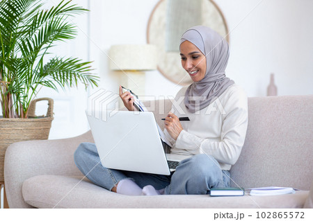 Young muslim business woman recruiter in hijab working remotely from home via laptop. Sits on the sofa and conducts online job interviews, conducts surveys and questionnaires, records, consults. 102865572
