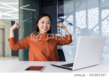 A young Asian business woman working in the office using a laptop, confidently and smilingly looking at the camera and showing a power gesture with her hands. Successfully completed work, project. A young Asian business woman working in the office using a laptop, confidently and smilingly looking at the camera and showing a power gesture with her hands. Successfully completed work, project. 102865587