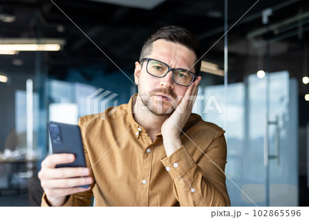 Upset young man sitting in the office holding the phone in his hands. Lost money in online games, received bad news via sms, holds his face worriedly and looks at the camera. Close-up photo. Upset young man sitting in the office holding the phone in his hands. Lost money in online games, received bad news via sms, holds his face worriedly and looks at the camera. Close-up photo. 102865596