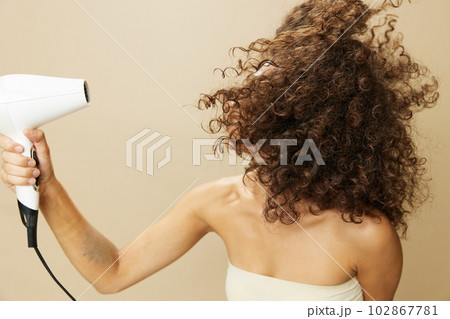 Woman dries curly afro hair with blow dryer, home beauty care styling products hair, smile on beige background Woman dries curly afro hair with blow dryer, home beauty care styling products hair, smile on beige background 102867781