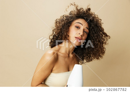 Woman dries curly afro hair with blow dryer, home beauty care styling products hair, smile on beige background Woman dries curly afro hair with blow dryer, home beauty care styling products hair, smile on beige background 102867782