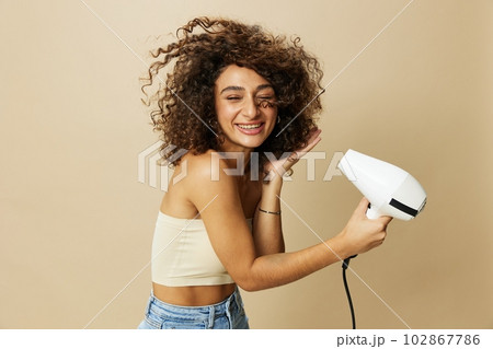 Woman dries curly afro hair with blow dryer, home beauty care styling products hair, smile on beige background 102867786