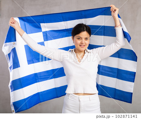 Middle-aged positive woman in casual clothes hold unfurled flag of Greece in hands raised above head Middle-aged positive woman in casual clothes hold unfurled flag of Greece in hands raised above head 102871141