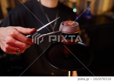 Burning coals in tongs are held in balance against the background of a burning blue burner. Burning coals in tongs are held in balance against the background of a burning blue burner. 102876510
