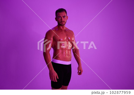 Man athletic body bodybuilder in briefs with nude torso abs full-length in the background fitness classes, purple colored light. Advertising, sports, active lifestyle, competition, challenge concept. Man athletic body bodybuilder in briefs with nude torso abs full-length in the background fitness classes, purple colored light. Advertising, sports, active lifestyle, competition, challenge concept. 102877959