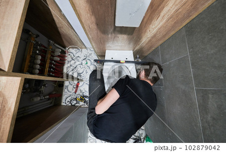 Top view of man installing flush toilet bowl in apartment under renovation. Male plumber crouching down on the floor with construction tools while working on toilet installation at home. 102879042