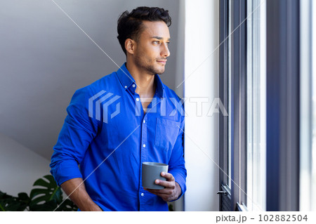 Happy biracial man holding mug and looking through window Happy biracial man holding mug and looking through window 102882504