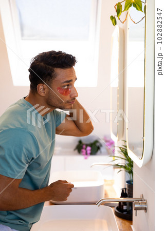 Biracial man looking in mirror and applying under eye masks in bathroom Biracial man looking in mirror and applying under eye masks in bathroom 102882547