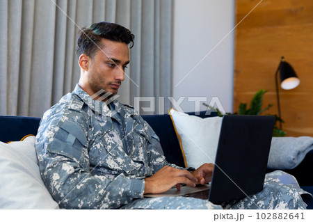 Biracial male soldier wearing military uniform, sitting on sofa using laptop at home Biracial male soldier wearing military uniform, sitting on sofa using laptop at home 102882641