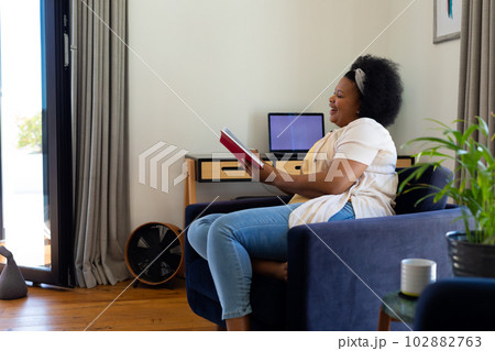Happy plus size african american woman sitting in armchair at home, reading book 102882763