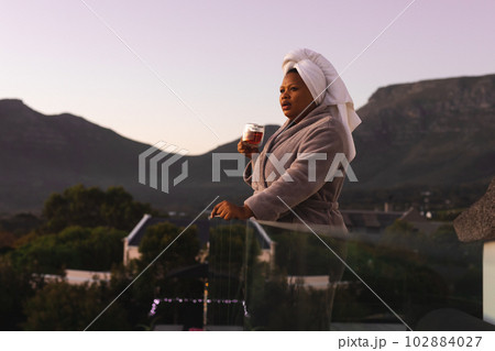 Happy plus size african american woman wearing robe and towel on head, drinking tea on balcony Happy plus size african american woman wearing robe and towel on head, drinking tea on balcony 102884027