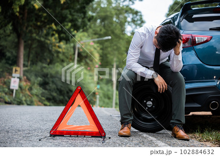 Young businessman and one mature man, sitting by a broken car with a warning red triangle sign in the background. The businessman is changing the wheel while the mature man looks worried. 102884632
