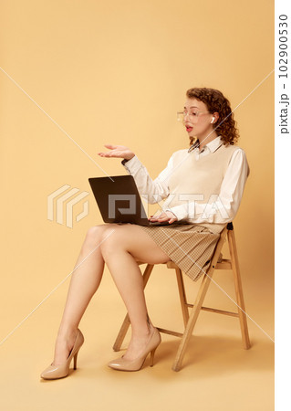 Portrait of young beautiful girl in formal clothes sitting on chair and having online video call on laptop against studio background Portrait of young beautiful girl in formal clothes sitting on chair and having online video call on laptop against studio background 102900530
