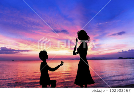 夕焼けに染まるビーチでプロポーズする男性と驚く女性のシルエット。の
