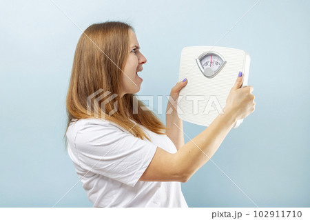 Young brunette woman looking at weight scale in shock on blue background Young brunette woman looking at weight scale in shock on blue background 102911710