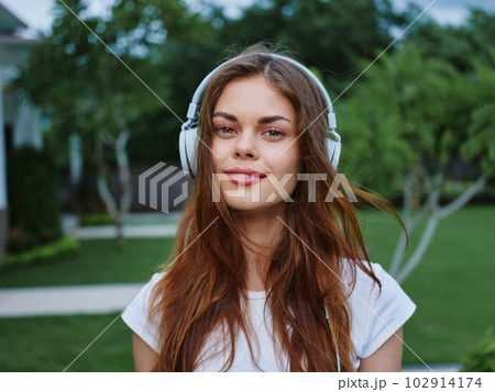 Woman wearing headphones listening to music smile with teeth looking toward camera in white t-shirt, traveling in Asia Woman wearing headphones listening to music smile with teeth looking toward camera in white t-shirt, traveling in Asia 102914174