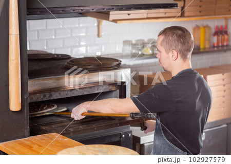 The chef prepares pizza. Raw pizza ready to bake. Cook in a blue apron in the kitchen. with a shovel in his hands. boxes for food delivery on background. 102927579