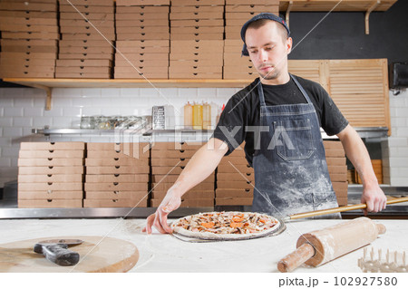 The chef prepares pizza. Raw pizza ready to bake. Cook in a blue apron in the kitchen. with a shovel in his hands. boxes for food delivery on background. The chef prepares pizza. Raw pizza ready to bake. Cook in a blue apron in the kitchen. with a shovel in his hands. boxes for food delivery on background. 102927580