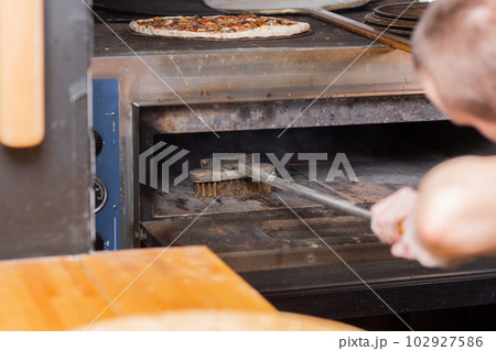 The chef cleaning the pizza oven. Raw pizza ready to bake. Cook in a blue apron in the kitchen. with a shovel in his hands. boxes for food delivery on background. 102927586