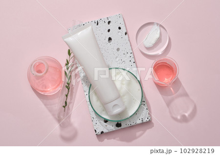 Top view of white plastic tube unlabeled container facial cleanser mockup for cosmetic on brick podium on pink background. Lab glassware containing pink liquid, cream texture and foam. 102928219