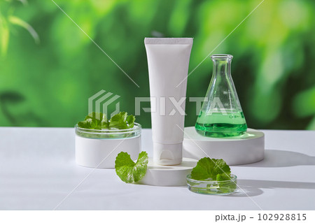 Unlabeled cosmetic container and an erlenmeyer flask of green liquid placed on round white podiums. Gotu kola (Centella asiatica) is widely used in skin care 102928815