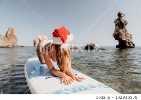 Woman sea sup. Close up portrait of happy young caucasian woman with long hair in Santa hat looking at camera and smiling. Cute woman portrait in a white bikini posing on sup board in the sea 102930839