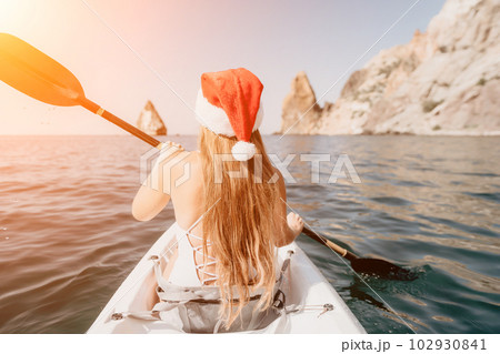 Woman in kayak back view. Happy young woman in Santa hat floating in kayak on calm sea. Summer holiday vacation and cheerful female people relaxing having fun on the boat. Woman in kayak back view. Happy young woman in Santa hat floating in kayak on calm sea. Summer holiday vacation and cheerful female people relaxing having fun on the boat. 102930841