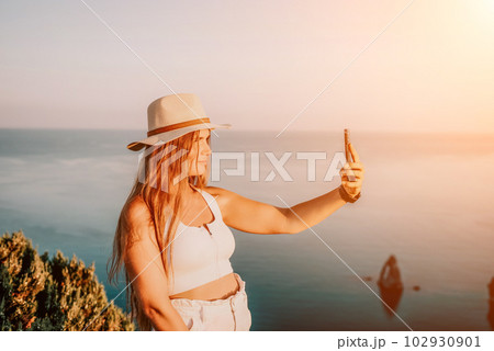 Woman travel sea. Happy tourist in hat enjoy taking picture outdoors for memories. Woman traveler posing on the beach at sea surrounded by volcanic mountains, sharing travel adventure journey. 102930901